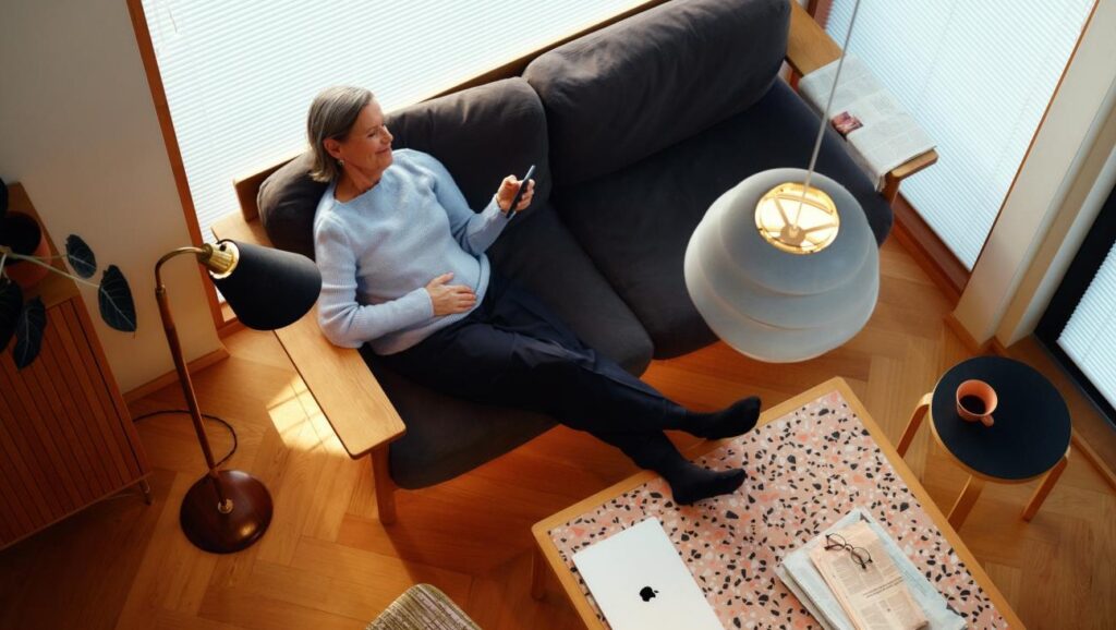 Kuvassa on keski-ikäinen nainen rentoutumassa modernissa olohuoneessa. Hän istuu tummalla sohvalla jalat ojennettuina ja pitää kädessään älypuhelinta, johon hän katsoo hymyillen. Naisella on päällään vaaleansininen pusero ja tummat housut. Sohvapöydällä, jossa on värikäs terrazzo-kuvioinen pinta, näkyy avattu kannettava tietokone, sanomalehti, silmälasit ja kahvikuppi. Tilassa on lämmin puulattia, design-tyylinen valaisin ja suuret ikkunat, joista tulvii luonnonvaloa.