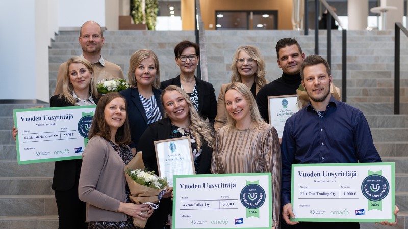 Ryhmä ihmisiä poseeraa portaikossa juhlallisessa tilaisuudessa. Etualalla kolme henkilöä pitelee "Vuoden Uusyrittäjä 2024" -palkintokylttejä, joissa näkyvät yritysten nimet ja summat: Lattiapalvelu Royal Oy (2 500 €), Akron Taika Oy (1 500 €) ja Flat Out Trading Oy (1 000 €). Heidän kanssaan on muita hymyileviä henkilöitä, joista osa pitää käsissään kukkakimppuja ja kunniakirjoja. Taustalla näkyy avoin, moderni sisätila ja portaikko. Ilmapiiri on iloinen ja juhlava.