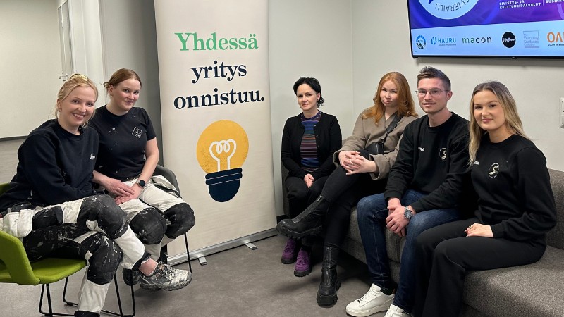 Kuusi nuorta aikuista istuu rennosti ryhmäkuvassa sisätiloissa. Kolme henkilöä istuu vihreillä tuoleilla ja kolme harmaalla sohvalla. Kaikilla on rennot vaatteet, osa pukeutunut mustiin työvaatteisiin ja maaliroiskeisiin housuihin. Keskellä on valkoinen pystybanneri, jossa lukee "Yhdessä yritys onnistuu" ja kuvassa on lamppu. Taustalla seinällä näkyy osittain näyttö, jossa on erilaisia logoja ja tekstejä. Tunnelma on iloinen ja rento.