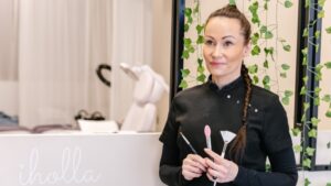 Hymyilevä kosmetologi seisoo opetushoitolassa meikkisiveltimet kädessään; taustalla hoitotuoli ja teksti "iholla"