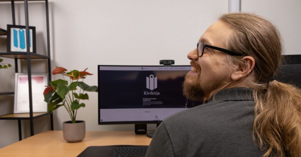 Tilitoimiston työntekijä toimistolla, kääntyy hymyillen tietokoneelta kameralle.